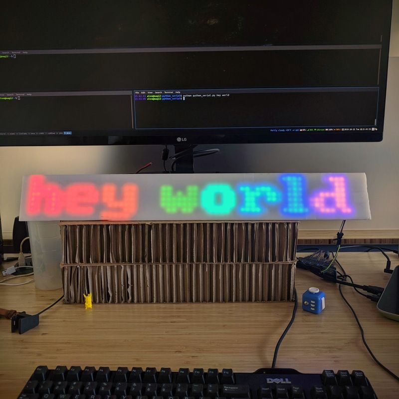 The words 'hey world' glow on a long acrylic rectangle under a computer monitor.